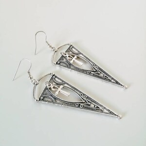 May include: Silver dangle earrings with a triangle shape and an ankh charm.