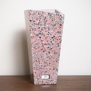 May include: A pink terrazzo vase with a rectangular shape and a slightly tapered design. The vase has a speckled pattern of black, white, and gray aggregate. A small, rectangular label with the letter "M" is affixed to the front.
