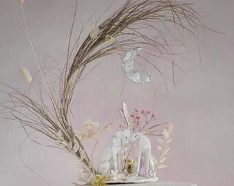 Op de afbeelding: Witte keramische konijnenfiguurtjes die elkaar kussen onder een zilveren halve maan, omringd door gedroogde bloemen en grassen op een witte taartstandaard.