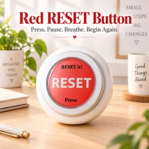 May include: A round white device featuring a large red circular button in the center. The button displays the text 'RESET it!', 'RESET', and 'Press' in black and white lettering. The device is positioned on a wooden surface.