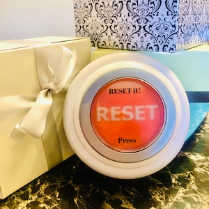 Op de afbeelding: Een witte en zilveren resetknop met het woord "RESET" in grote, witte letters op een rode achtergrond. De tekst "RESET it!" staat boven het woord "RESET", en "Press" staat eronder. De knop is omgeven door een wit en zilverkleurig frame. Cadeaudozen staan op de achtergrond.