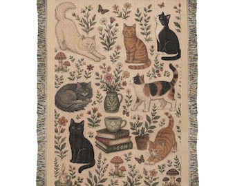 May include: A woven throw blanket featuring a beige background with a pattern of various cats, including a stretching cream cat, a tabby, a black and white cat, a curled gray cat, a calico, and a black cat, surrounded by flowers, mushrooms, and books.