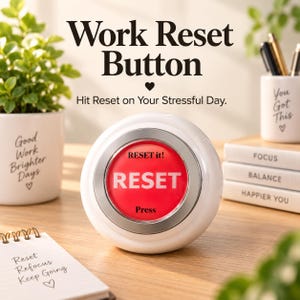 May include: A round white desk accessory featuring a large red circular button in the center with the word RESET printed in white, along with the text RESET it! and Press. The device is surrounded by office items including a plant and a notepad.