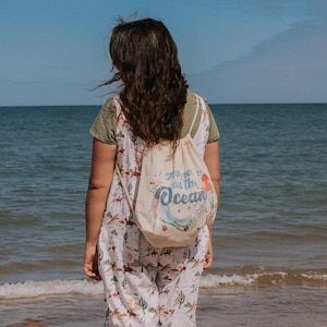 May include: A person standing on a sandy beach, facing the ocean. They are wearing a white and green floral jumpsuit and a white drawstring bag with the text "go with the ocean" printed on it.