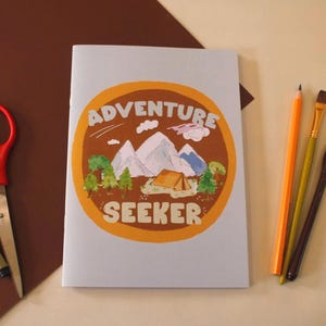 May include: A light gray notebook featuring a circular orange emblem with a mountain landscape, a tent, and green trees. The text ADVENTURE SEEKER is printed in white. Beside the notebook are red scissors, a black pen, an orange pencil, and two brushes.