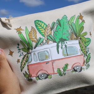May include: A white and pink canvas zippered pouch with a hand-painted design of a vintage van with green plants growing out of the roof. The van is surrounded by gold stars.