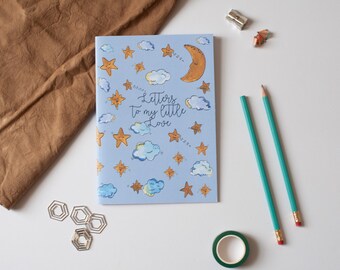 May include: A blue notebook with a whimsical design featuring stars, clouds, and a crescent moon. The text "Letters to my little Love" is written in a cursive font.