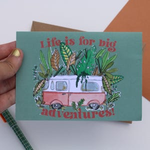 May include: A teal greeting card with a red and white camper van illustration, surrounded by green and brown leaves. The text "Life is for big adventures!" is written in red.