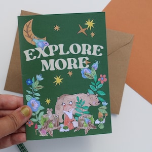 May include: A green greeting card with the words "EXPLORE MORE" in white. The card features illustrations of a bear, fox, and other forest elements. A brown envelope is behind the card.