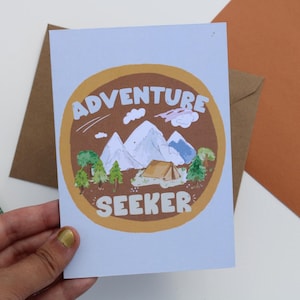 May include: A greeting card with a brown and white illustration of mountains, trees, and a tent inside a brown circle. The words "ADVENTURE SEEKER" are written in white. A brown envelope is behind the card.