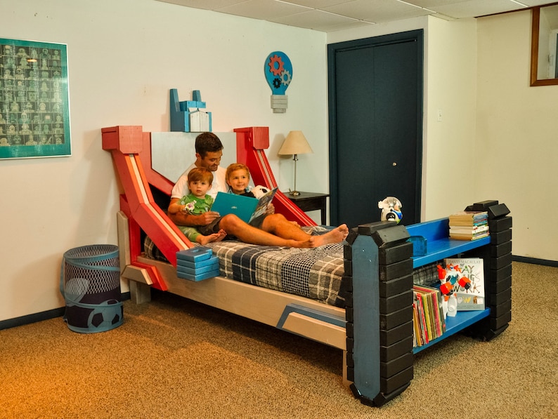 May include: A red, blue, and gray bed frame with a built-in bookshelf. A father and two children are sitting on the bed, reading a book. The bed frame has a unique design with a robot-like appearance.