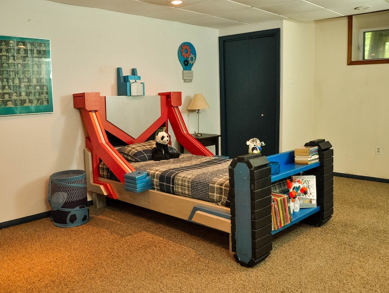 May include: A red and blue robot-themed bed frame with a plaid comforter and a panda bear plush toy. The bed has a built-in bookshelf with blue shelves and a black and white graphic on the side.