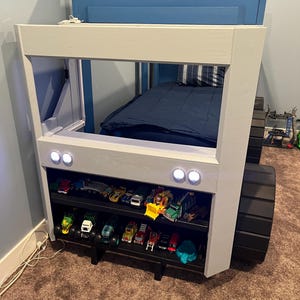 Puede incluir: Una estructura de cama de madera blanca y azul con forma de cami&oacute;n con ruedas negras. La cama tiene un colch&oacute;n azul y una cabecera blanca con dos peque&ntilde;as luces blancas. La cama tiene dos estantes debajo para guardar juguetes. Los estantes est&aacute;n llenos de coches de juguete.