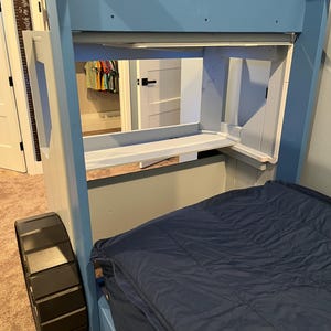 Puede incluir: Una litera de madera azul con una rueda de neum&aacute;tico negra en la litera inferior. La litera superior tiene un estante y una peque&ntilde;a abertura para almacenamiento. La cama est&aacute; hecha con un edred&oacute;n azul.