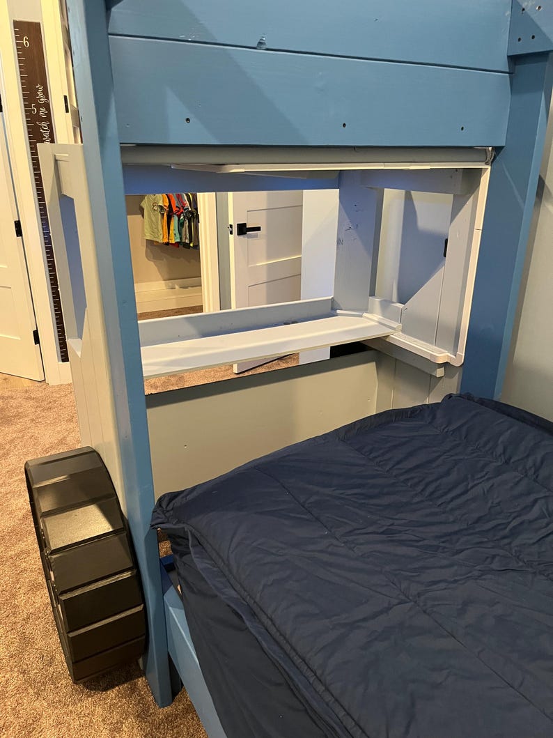Puede incluir: Una litera de madera azul con una rueda de neum&aacute;tico negra en la litera inferior. La litera superior tiene un estante y una peque&ntilde;a abertura para almacenamiento. La cama est&aacute; hecha con un edred&oacute;n azul.