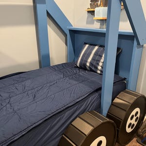 Puede incluir: Una estructura de cama de madera azul con un colch&oacute;n azul y ruedas negras. La estructura de la cama tiene forma de casa con techo y ventanas. Las ruedas tienen detalles blancos y radios negros.