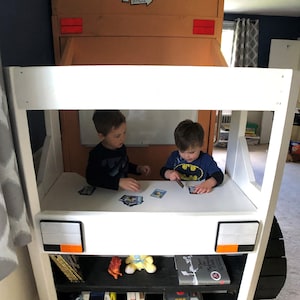 May include: Two children playing with cards at a white table inside a white and brown toy truck bed. The truck bed has a black shelf with books and toys. The truck has the words "Schmidt Bros." on the side.