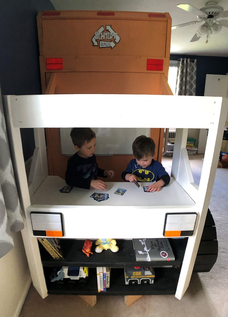May include: Two children playing with cards at a white table inside a white and brown toy truck bed. The truck bed has a black shelf with books and toys. The truck has the words "Schmidt Bros." on the side.