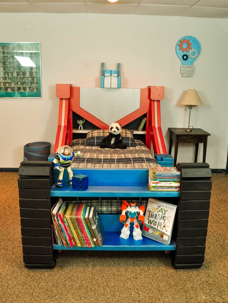 May include: A red, blue, and black wooden bed frame with a built-in bookshelf. The bed frame is decorated with a robot theme and features a panda bear plush toy, a robot toy, and a book titled "The Way Things Work Now".