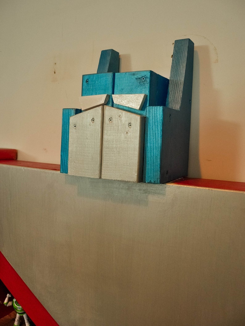 May include: A wooden toy robot head painted in shades of blue and silver. The robot head is mounted on a wall.