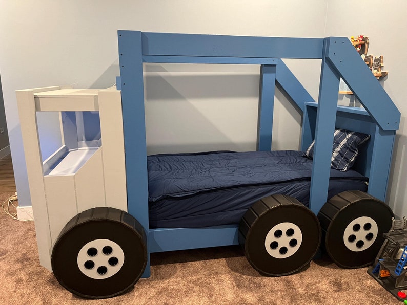 Puede incluir: Una cama de cami&oacute;n de madera azul y blanca con un colch&oacute;n azul y ruedas negras. La cama tiene una escalera en el lateral y un estante en la parte trasera.