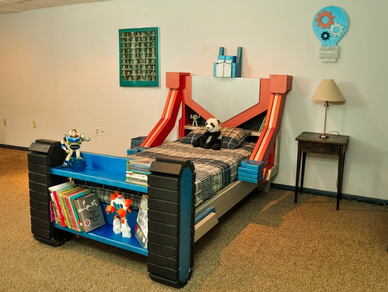 May include: A red, blue, and white themed bed frame with a built-in bookshelf. The bed frame is decorated with a robot design. A panda bear plush toy is sitting on the bed.