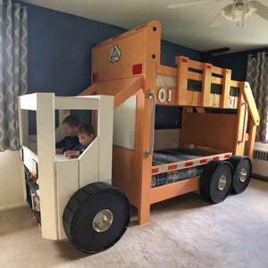May include: A wooden bunk bed shaped like a dump truck with a white cab and a brown bed. Two children are sitting in the cab of the truck. The bed has a black and white checkered mattress and black tires.