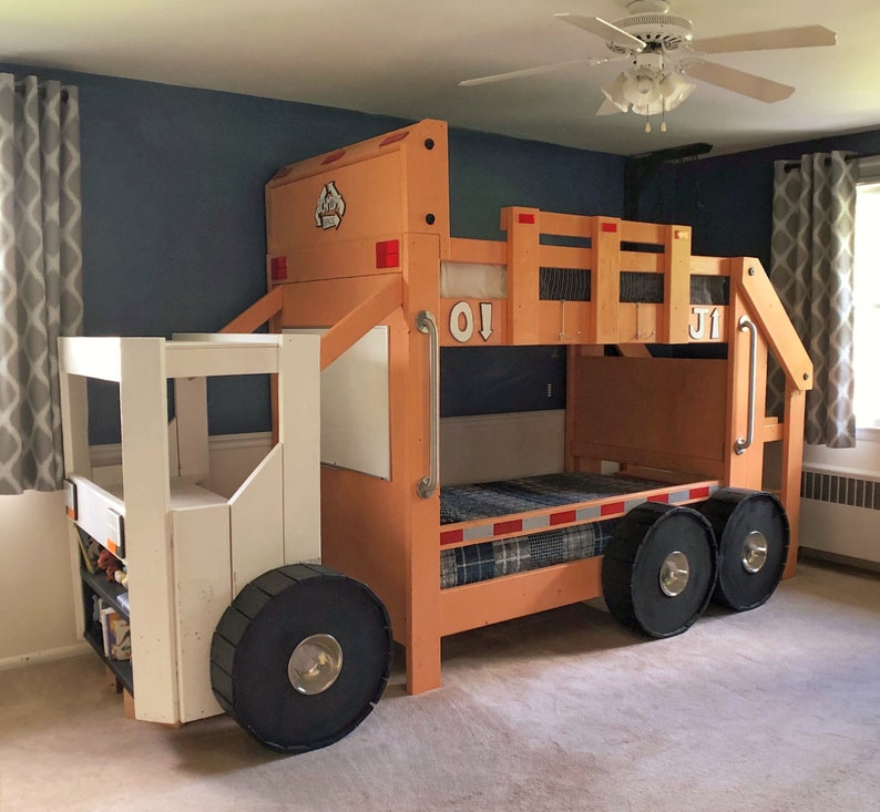 May include: A wooden bunk bed shaped like a toy truck with a white cab and orange bed frame. The bed has a ladder on the side and a small shelf in the cab. The bed is decorated with the numbers "01" and "J1" on the side.