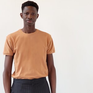 Op de afbeelding: Een man draagt een lichtbruin t-shirt met een ronde hals en een zwarte pantalon met pinstripes.
