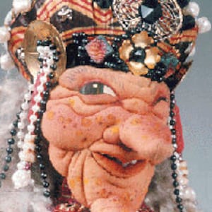 Puede incluir: Primer plano de la cara de una muñeca hecha a mano con una nariz grande y bulbosa, una amplia sonrisa con dientes y un solo ojo grande y verde. La muñeca lleva un cubrecabezas colorido y estampado con cuentas y otros adornos.