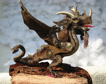 Puede incluir: Un títere de dragón marrón y dorado con una cola larga y alas. El dragón tiene una corona en la cabeza y la lengua roja sobresaliendo. El dragón está sentado sobre una superficie de tela marrón.