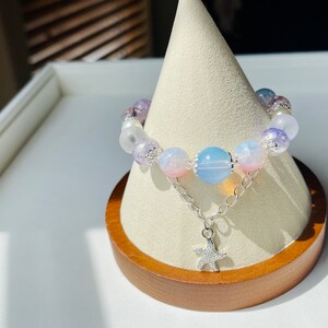 May include: A beaded bracelet featuring a mix of pastel-colored beads, including pink, blue, and purple, strung together with a silver chain and a starfish charm. The bracelet is displayed on a wooden stand.