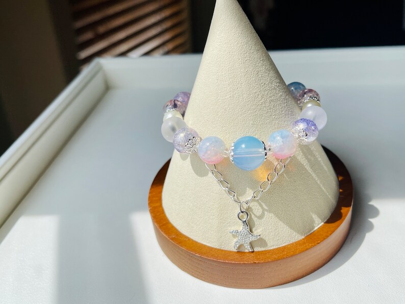 May include: A beaded bracelet featuring a mix of pastel-colored beads, including pink, blue, and purple, strung together with a silver chain and a starfish charm. The bracelet is displayed on a wooden stand.