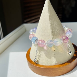 May include: A colorful beaded bracelet with a variety of pastel-colored beads, including pink, blue, and clear, is displayed on a white stand. The bracelet has silver-colored accents and dangling heart charms.