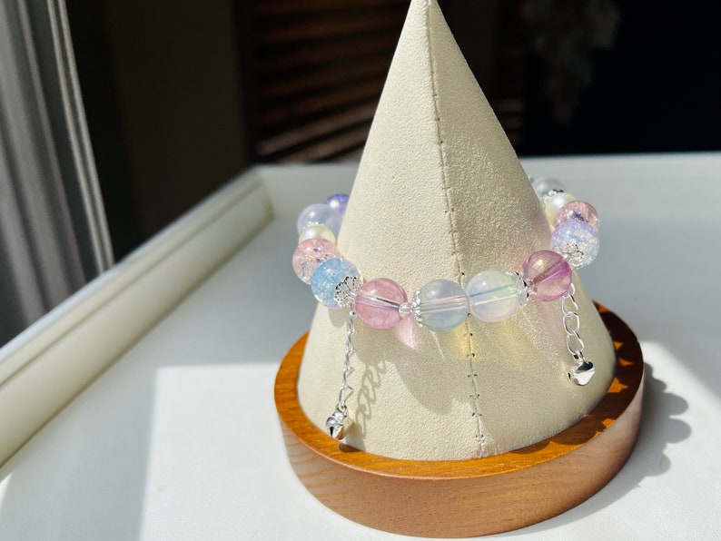 May include: A colorful beaded bracelet with a variety of pastel-colored beads, including pink, blue, and clear, is displayed on a white stand. The bracelet has silver-colored accents and dangling heart charms.