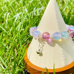 May include: A beaded bracelet with iridescent, pastel-colored beads in shades of pink, blue, and purple. The bracelet has silver-colored charms with heart-shaped pendants. The bracelet is displayed on a white cone-shaped stand.