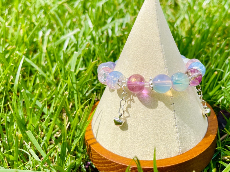 May include: A beaded bracelet with iridescent, pastel-colored beads in shades of pink, blue, and purple. The bracelet has silver-colored charms with heart-shaped pendants. The bracelet is displayed on a white cone-shaped stand.