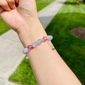 May include: A beaded bracelet featuring a mix of pink, purple, and iridescent beads. The bracelet has a silver chain with a small heart charm. The beads are various sizes and shapes, creating a unique and eye-catching accessory.
