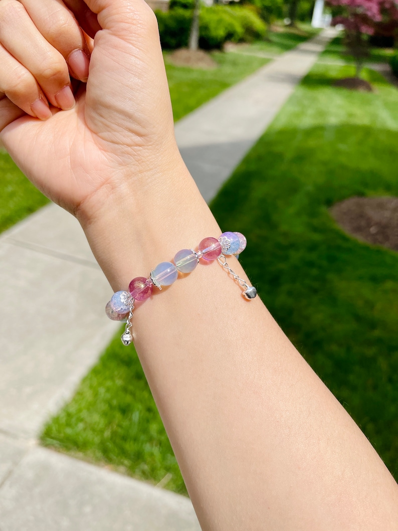 May include: A beaded bracelet featuring a mix of pink, purple, and iridescent beads. The bracelet has a silver chain with a small heart charm. The beads are various sizes and shapes, creating a unique and eye-catching accessory.