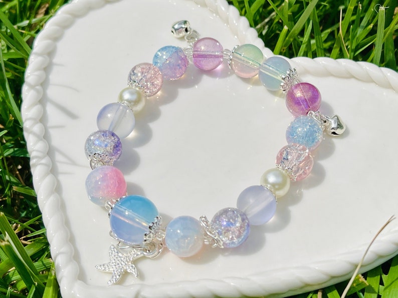 May include: A pastel-colored beaded bracelet with a mix of round and pearl-shaped beads. The bracelet features silver accents, small bells, and a starfish charm. The beads are in shades of pink, blue, purple, and white.