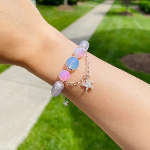May include: A beaded bracelet featuring a mix of pastel-colored beads, including pink, blue, and iridescent elements. A silver chain with a starfish charm dangles from the bracelet. The bracelet is worn on a wrist.