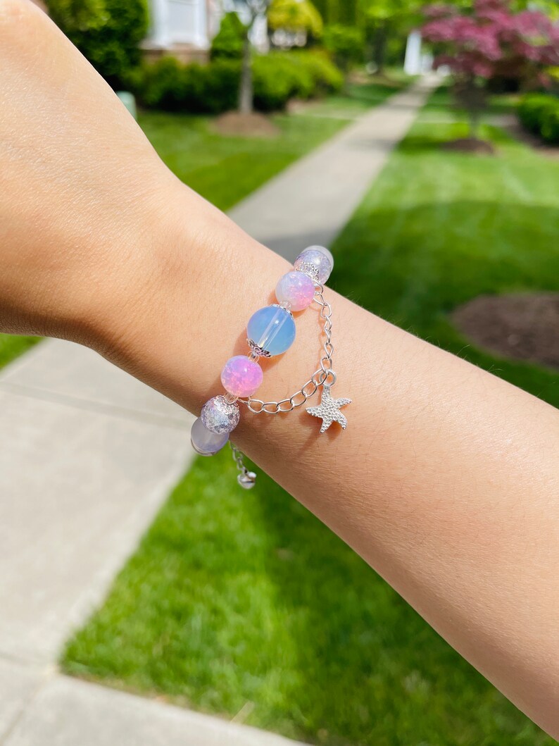 May include: A beaded bracelet featuring a mix of pastel-colored beads, including pink, blue, and iridescent elements. A silver chain with a starfish charm dangles from the bracelet. The bracelet is worn on a wrist.
