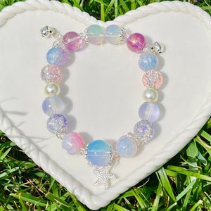 May include: A beaded bracelet featuring a variety of pastel-colored beads, including pink, blue, purple, and iridescent spheres. The bracelet has a silver starfish charm and small silver bells at each end. The bracelet is displayed on a white heart-shaped plate.