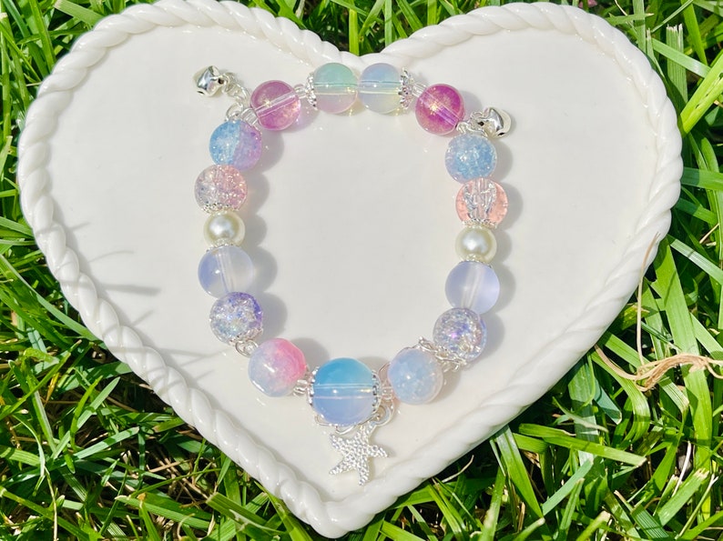 May include: A beaded bracelet featuring a variety of pastel-colored beads, including pink, blue, purple, and iridescent spheres. The bracelet has a silver starfish charm and small silver bells at each end. The bracelet is displayed on a white heart-shaped plate.