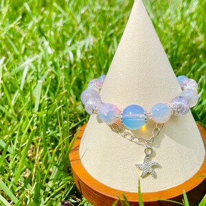 May include: A beaded bracelet with a starfish charm. The bracelet features a mix of light blue, pink, and clear beads. A silver chain connects the beads to a starfish pendant. The bracelet is displayed on a white cone-shaped stand.