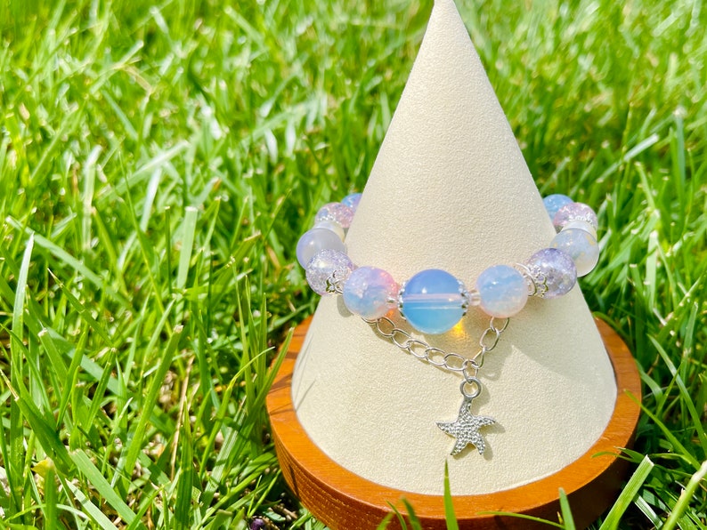 May include: A beaded bracelet with a starfish charm. The bracelet features a mix of light blue, pink, and clear beads. A silver chain connects the beads to a starfish pendant. The bracelet is displayed on a white cone-shaped stand.