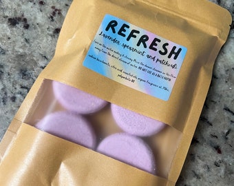 May include: A brown paper bag with a clear window containing four lavender-colored shower steamers. The bag has a label that reads "REFRESH Lavender spearmint and patchouli". The label also includes instructions and ingredients.