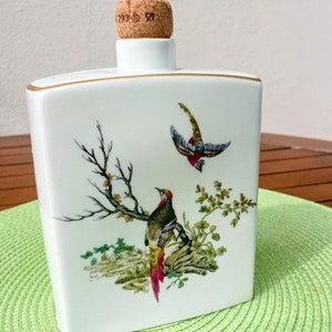 Op de afbeelding: Witte keramische fles met een kurken stopper, gouden rand en een geschilderd fazantenontwerp. Het kunstwerk bevat een zittende fazant en een andere in vlucht, met takken en bladeren. De kurk heeft de nummers "293" en "521" erop.