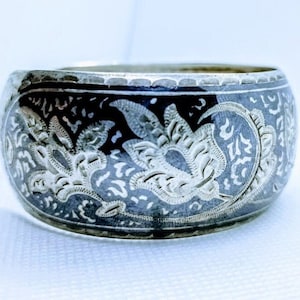 Könnte beinhalten: Ein silberfarbener Metall-Serviettenring mit einem eingravierten Blumendesign. Der Ring hat einen dunklen Hintergrund und ein detailliertes, erhabenes Blumenmuster. Das Design umschließt den gesamten Ring und erzeugt einen dekorativen und eleganten Look.