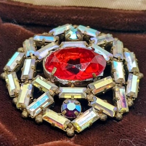 Peut inclure: Broche vintage avec une grosse pierre centrale rouge facettée, entourée de cristaux rectangulaires irisés. La broche a un sertissage en métal doré et une petite pierre ronde irisée. Un bijou classique.